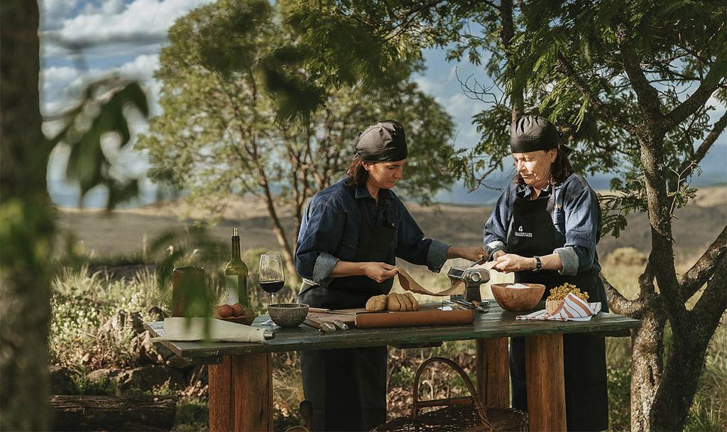 Dos cocineras preparando empanadas en una mesa al aire libre, montañas y árboles nativos de fondo